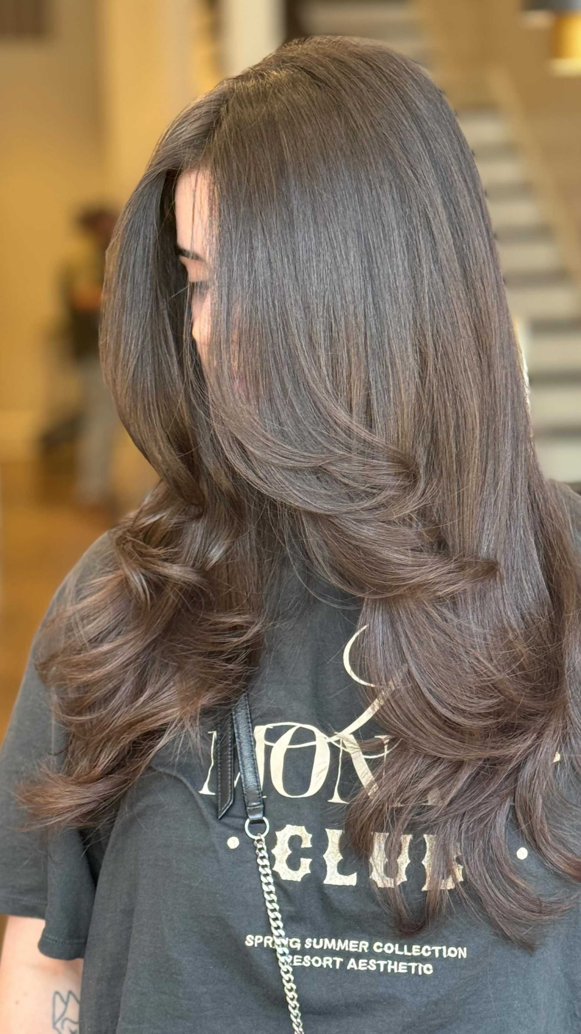 Woman with long, wavy brown hair and a casual t-shirt in a bright indoor setting.